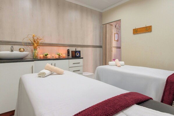 Habitación de hotel con dos camas, un lavamanos, un espejo y una pequeña mesa con un jarrón de flores secas.