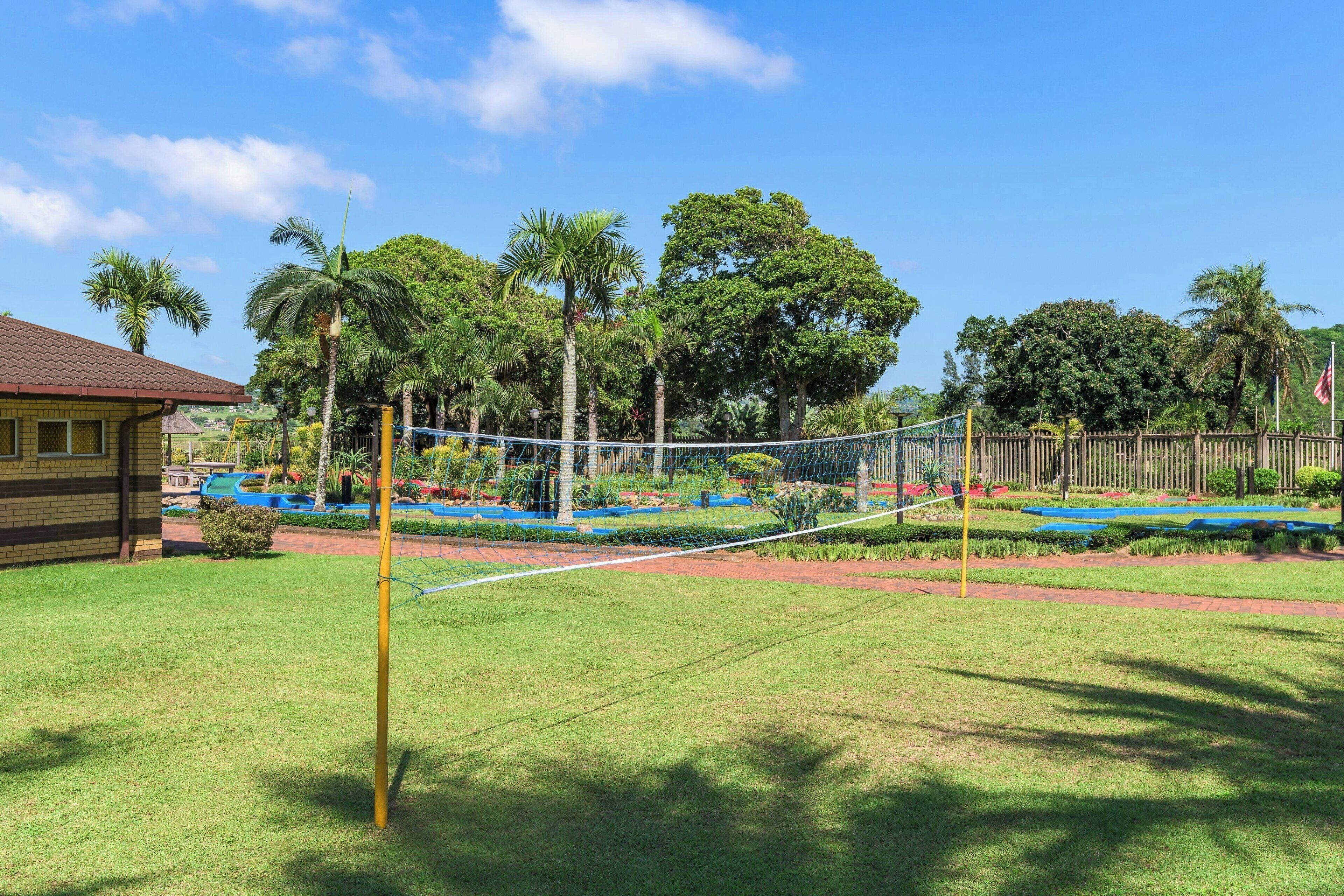 En legeplads med en volleyballbane med net, omgivet af frodig vegetation og palmer.