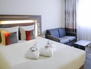 A hotel room with a bed, a sofa, a small table, and a nightstand with a lamp. - Novotel Lugano Paradiso (Paradiso)