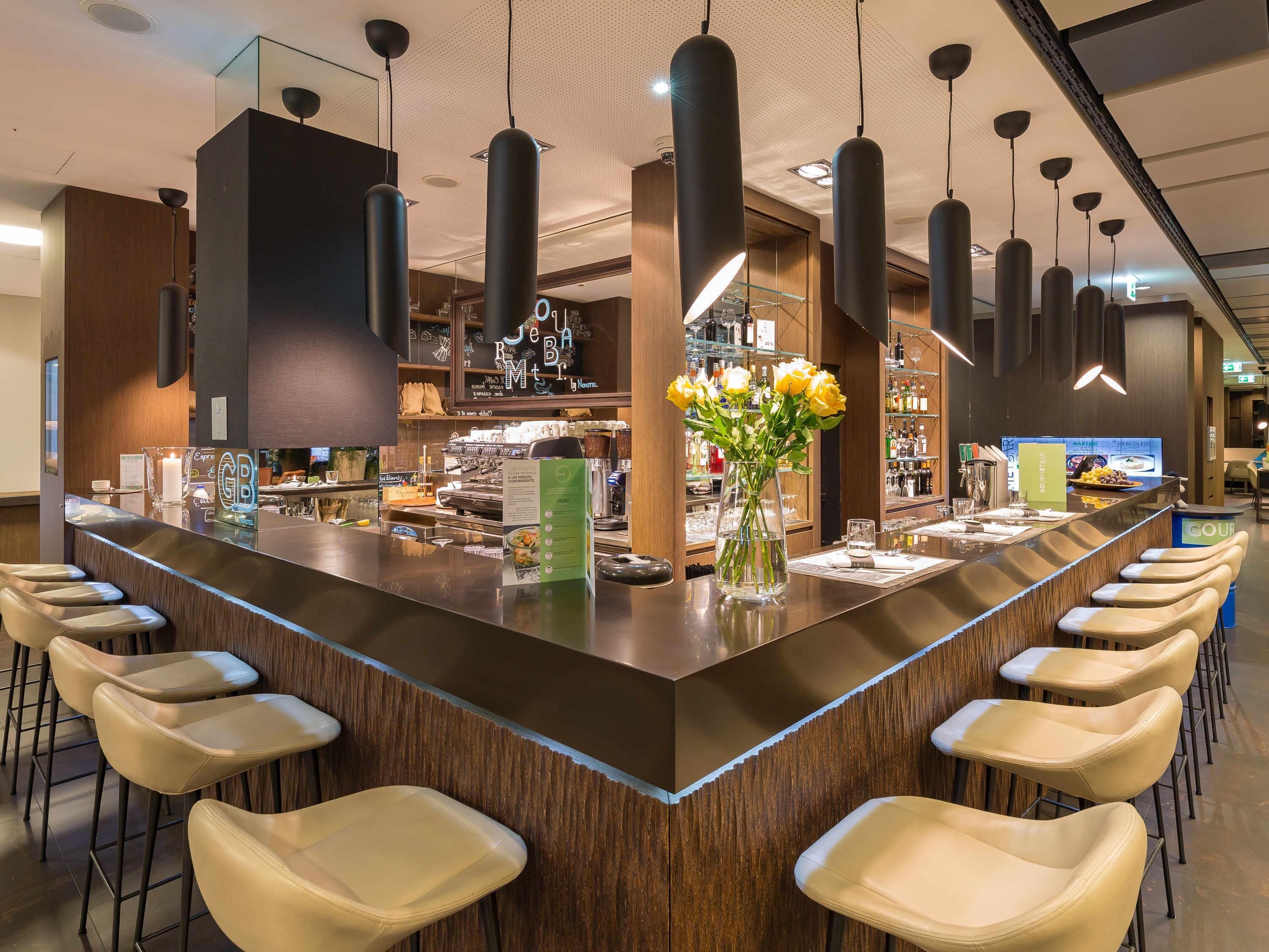 A modern bar area with a long counter, multiple hanging pendant lights, and a vase of yellow flowers.