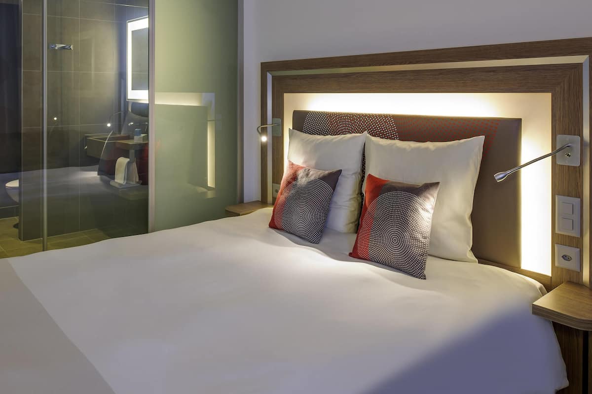 Een moderne hotelkamer met een douchecabine van glas, een houten hoofdeinde en een keurig opgemaakt bed.