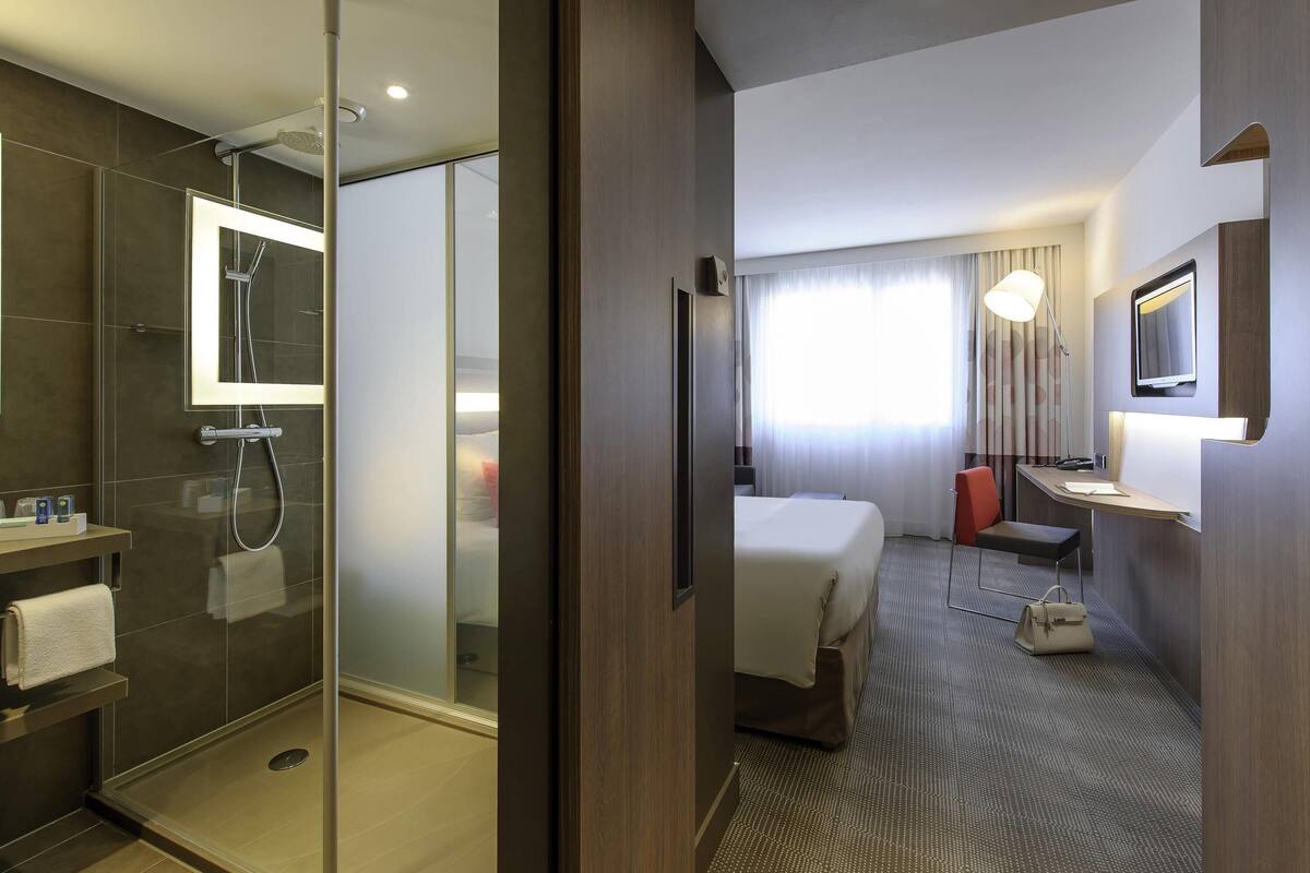 Hotelkamer met een douchecabine van glas, een bed, een bureau, een stoel, een nachtkastje en een spiegel.