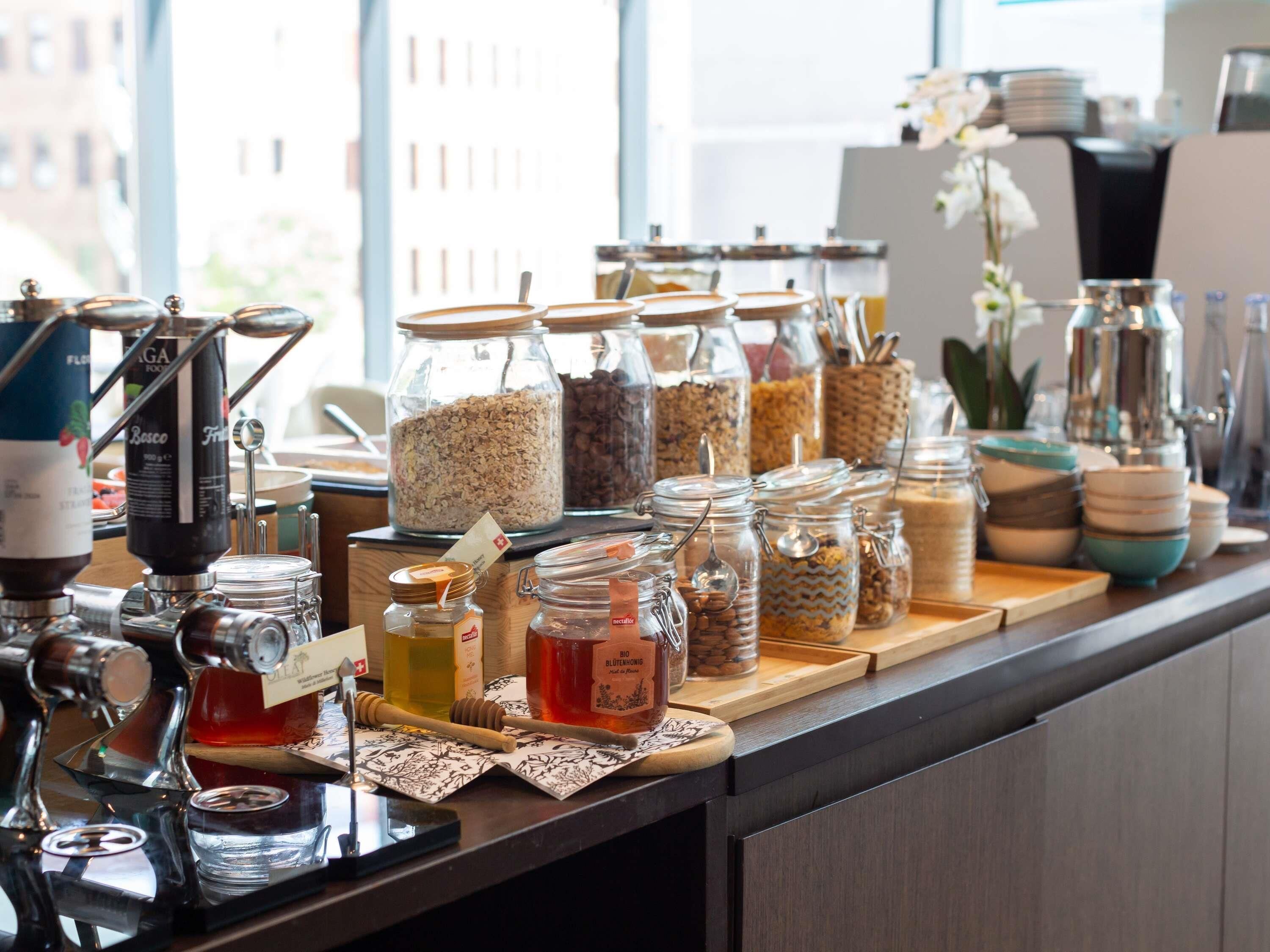 Una colazione a buffet con diversi cibi e condimenti, tra cui barattoli di granola e miele, e una macchina per il caffè.