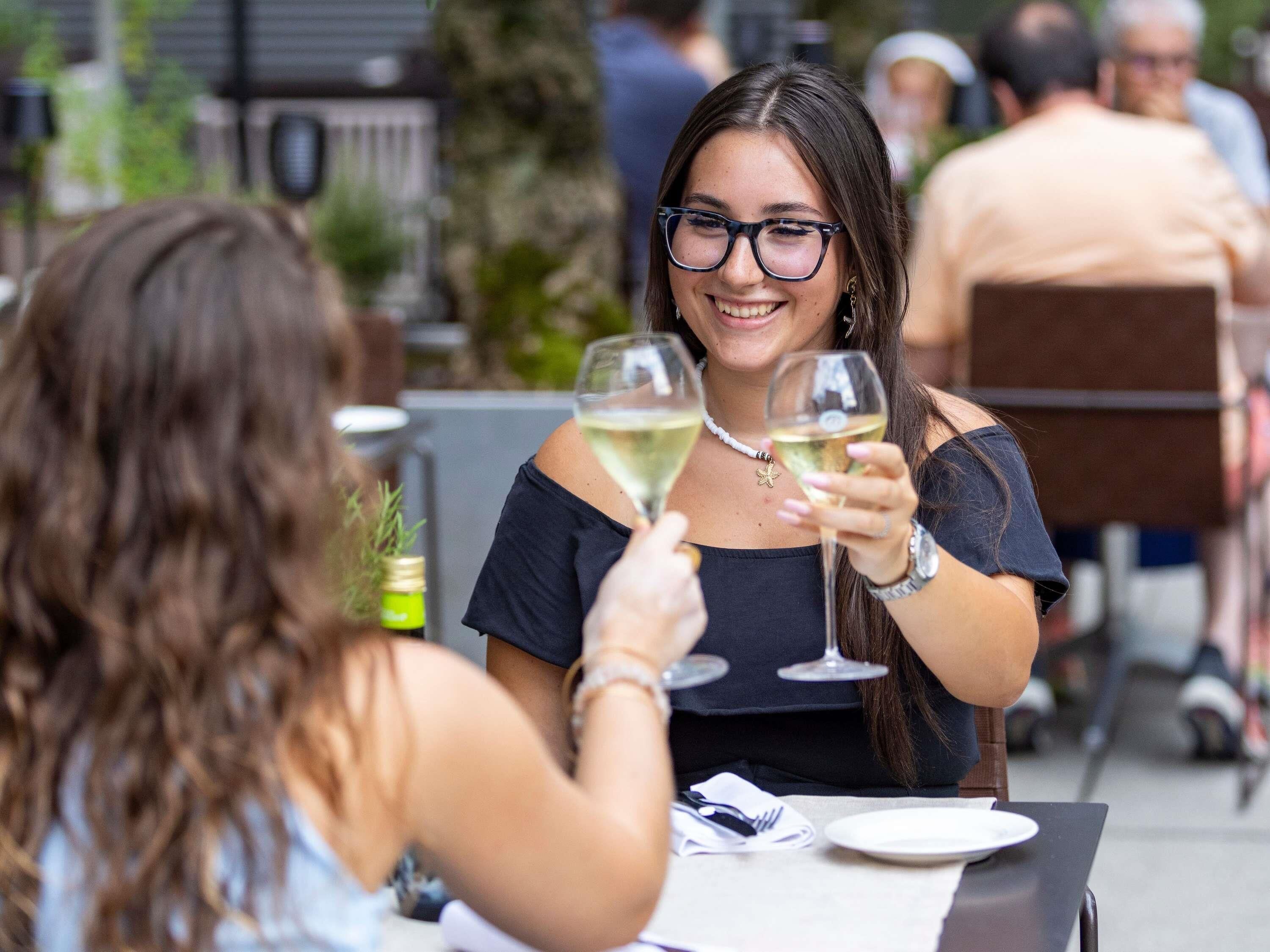 Zwei Personen stoßen mit Weingläsern an einem Tisch im Freien an. Eine weiße Platte und eine Flasche Olivenöl sind sichtbar.