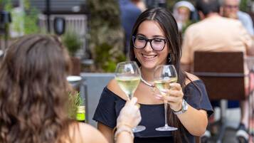 Zwei Personen stoßen mit Weingläsern an einem Tisch im Freien an. Eine weiße Platte und eine Flasche Olivenöl sind sichtbar.