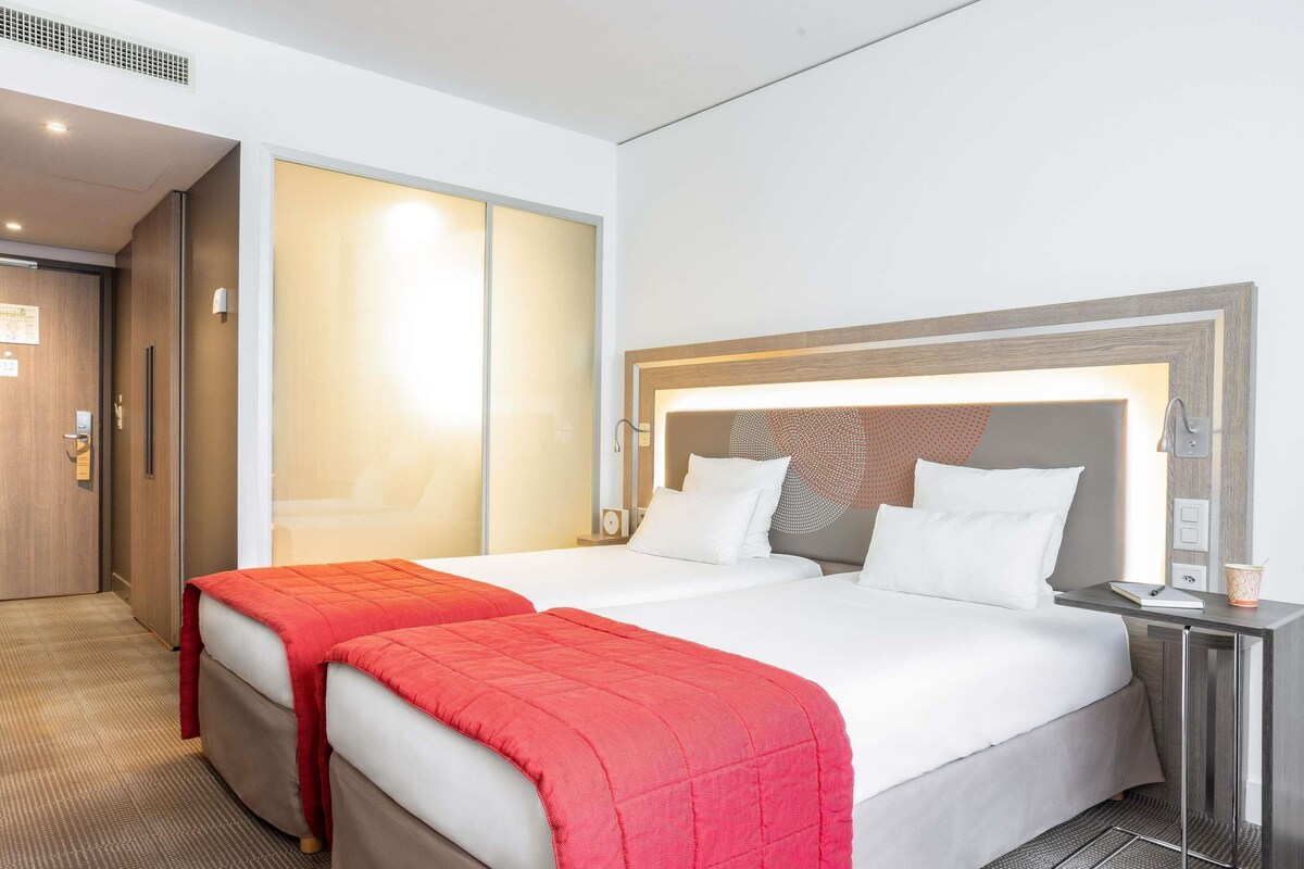Habitación de hotel con dos camas, cabecero de madera, mesita de noche con lámpara y puerta que da a un armario.