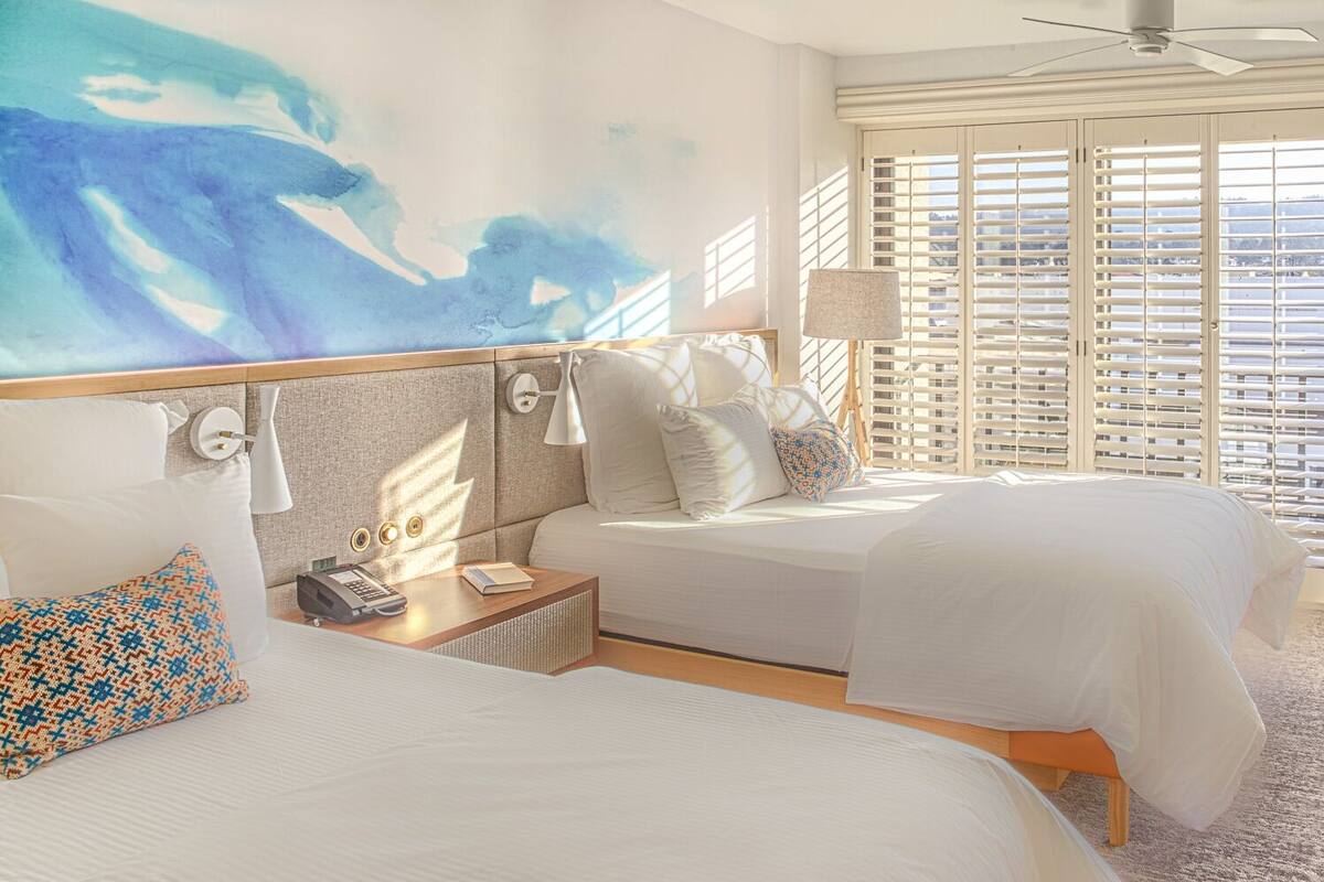 A hotel room with two beds, a large window with blinds, a ceiling fan, and a small table with a chair.