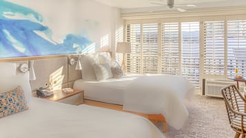 A hotel room with two beds, a large window with blinds, a ceiling fan, and a small table with a chair.