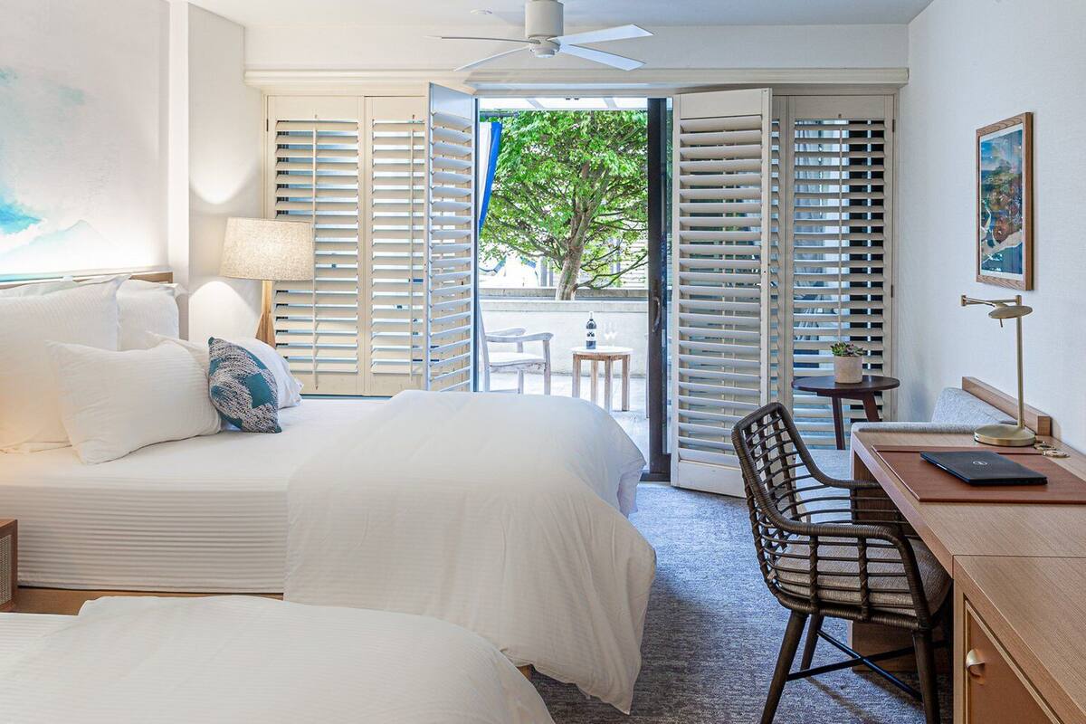 A hotel room with two beds, a desk, a chair, a TV, and a view of the outdoors.