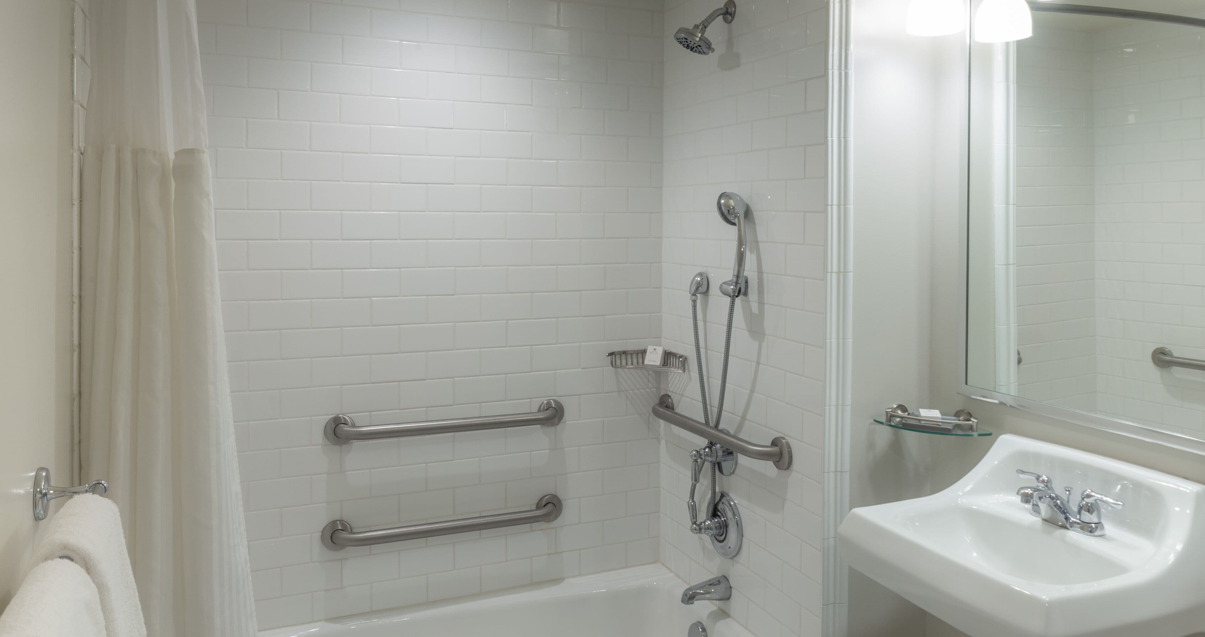 A bathroom with white tiled walls, a bathtub, a shower with grab bars, and a sink with a mirror.