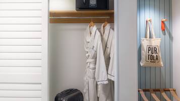 A hotel room with a wooden shelf, a microwave, a hanging robe, a suitcase, and a 'Pura Vida' tote bag.