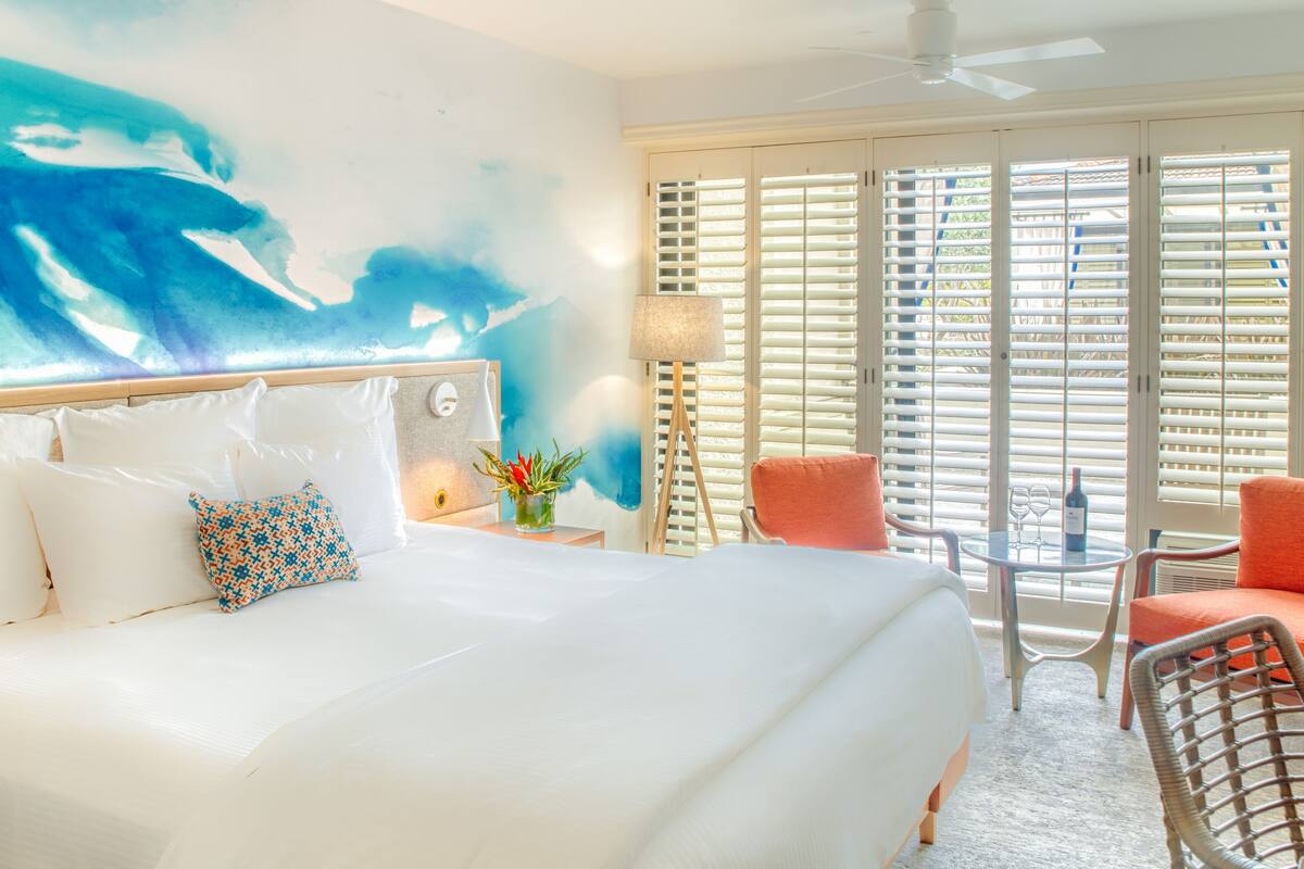 A hotel room with a large bed, a desk with two chairs, a window with blinds, and a ceiling fan.