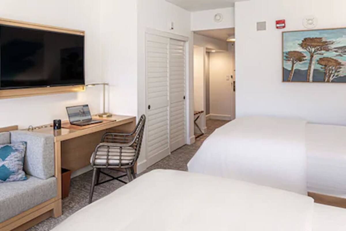 A hotel room with a bed, a desk with a laptop, a chair, a TV, and a painting on the wall.