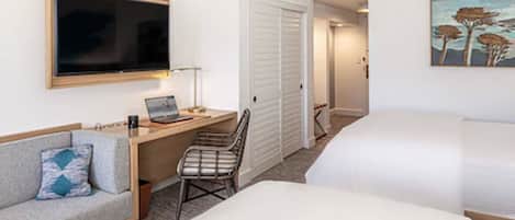A hotel room with a bed, a desk with a laptop, a chair, a TV, and a painting on the wall.