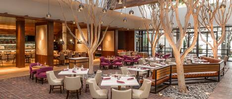 A spacious restaurant with high ceilings, exposed wooden beams, and hanging spherical lights. Tables are arranged around a central seating area with a long bench.