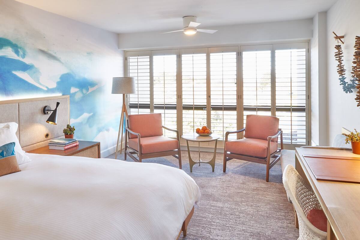 A hotel room with a large bed, two armchairs, a small table with fruit, a lamp, and a view through large windows.