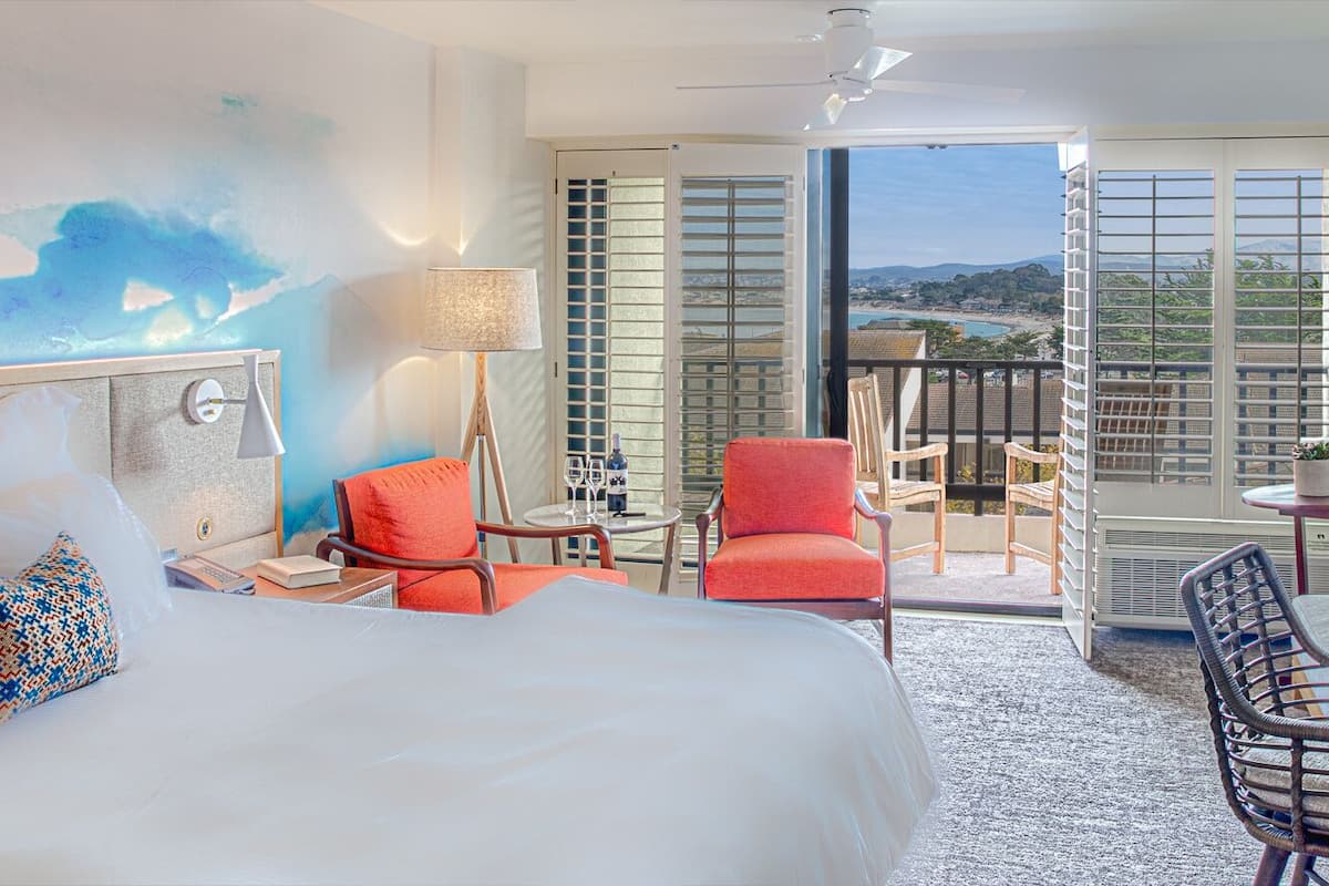 A hotel room with a bed, a desk, a chair, a balcony with a view, and a painting on the wall.