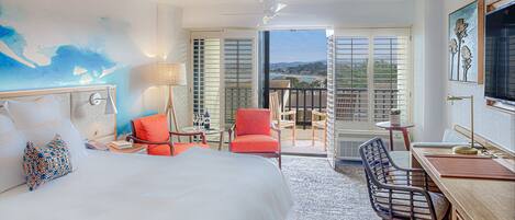 A hotel room with a bed, a desk, a chair, a balcony with a view, and a painting on the wall.