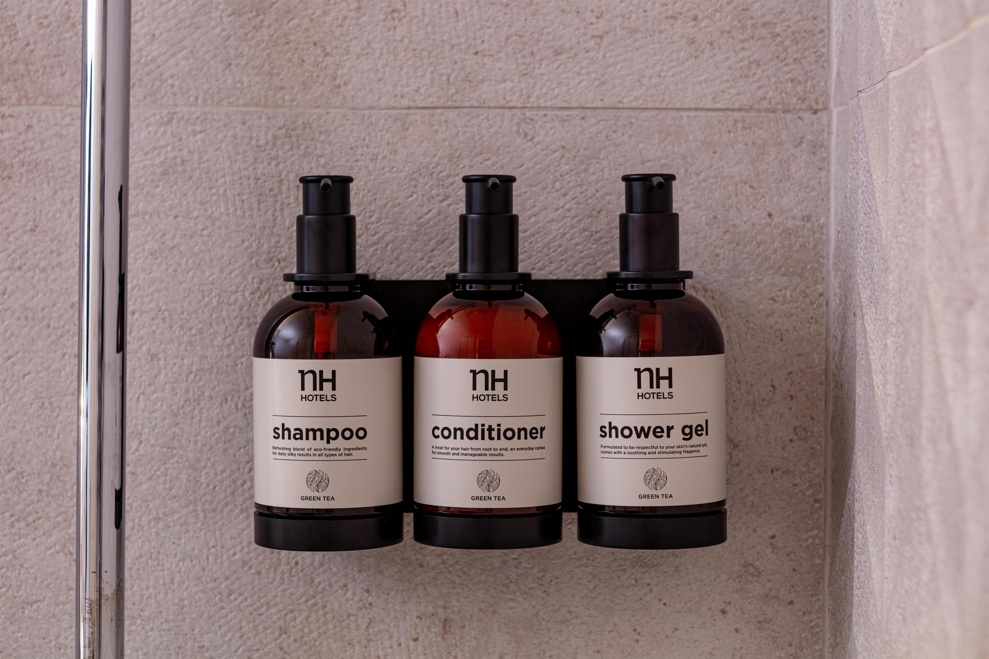 Trois flacons de produits d’accueil offerts par l’hôtel : shampoing, après-shampoing et gel douche, tous portant la mention « nh hotels » et « thé vert ».