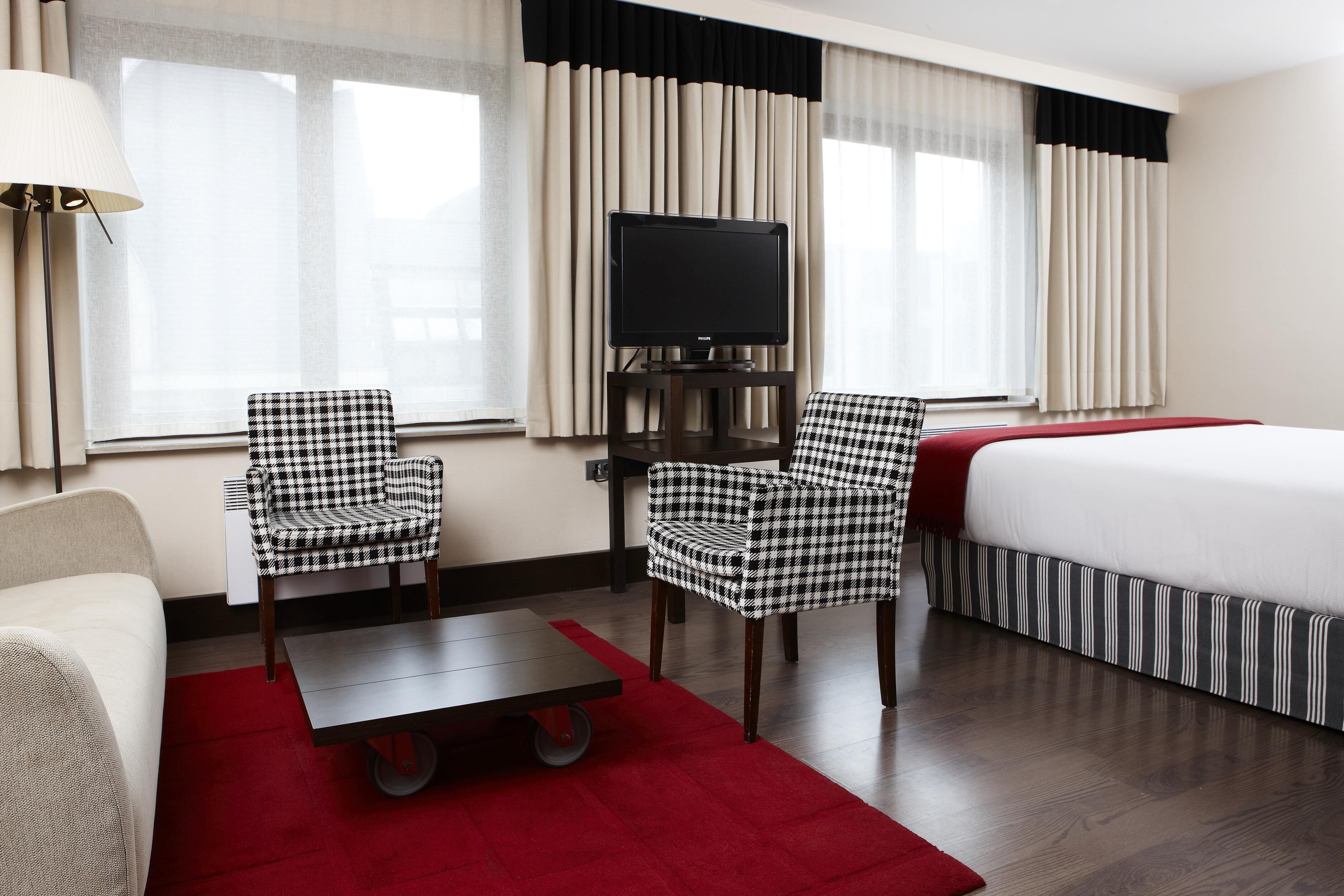A hotel room with a bed, two checkered chairs, a small table, a TV on a stand, and a red rug.
