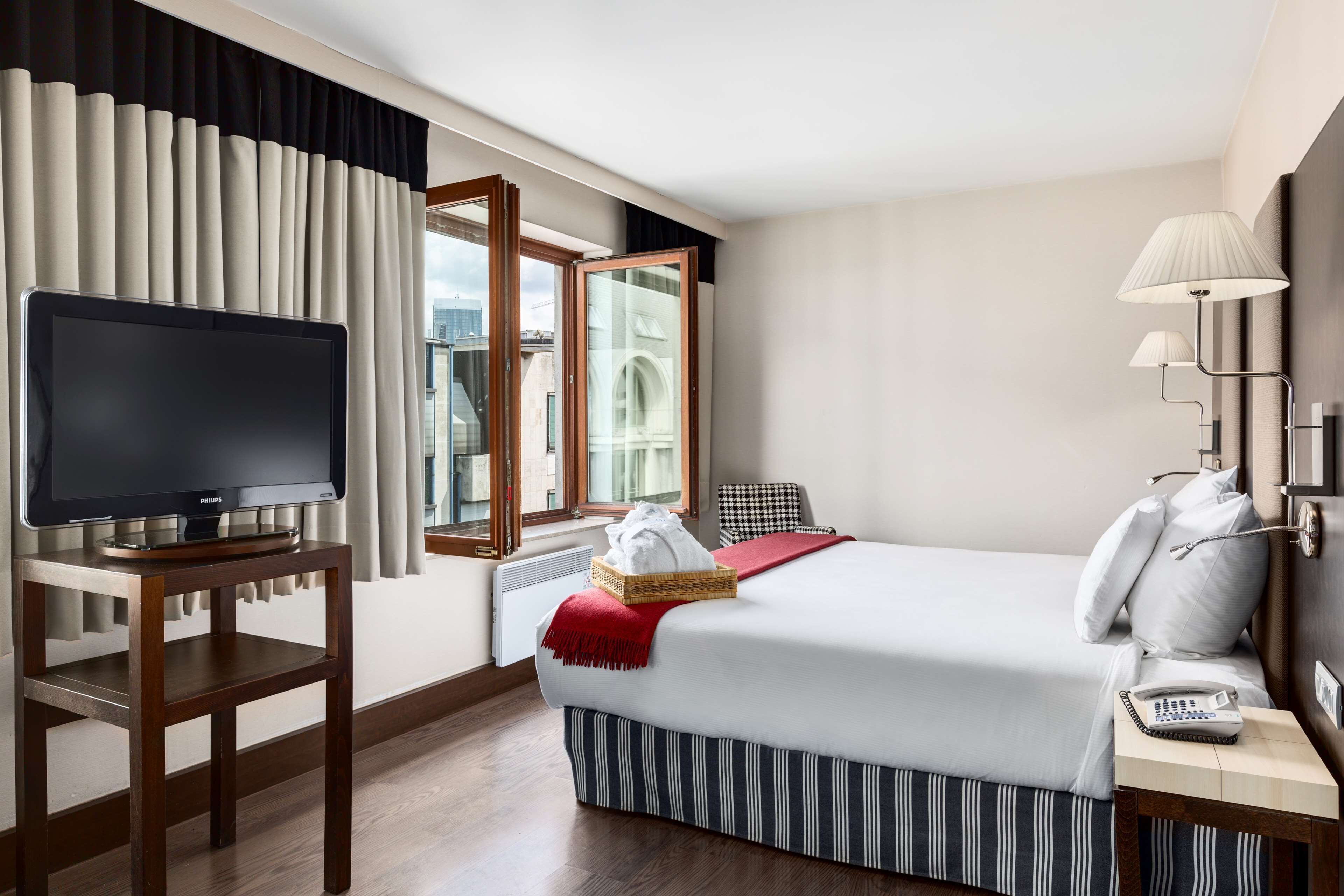 Une chambre d’hôtel avec un lit, un téléviseur posé sur une table en bois, une lampe et une vue sur un immeuble par la fenêtre.