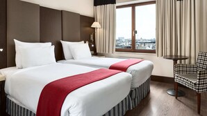 A hotel room with two beds, a view of the city, a chair, and a lamp. - NH Brussels Grand Place Arenberg (Brussels)