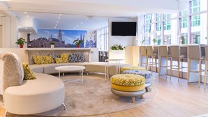 A modern hotel lobby with a curved sofa, ottomans, a small table, and a bar area with chairs. - NH Brussels Grand Place Arenberg (Brussels)