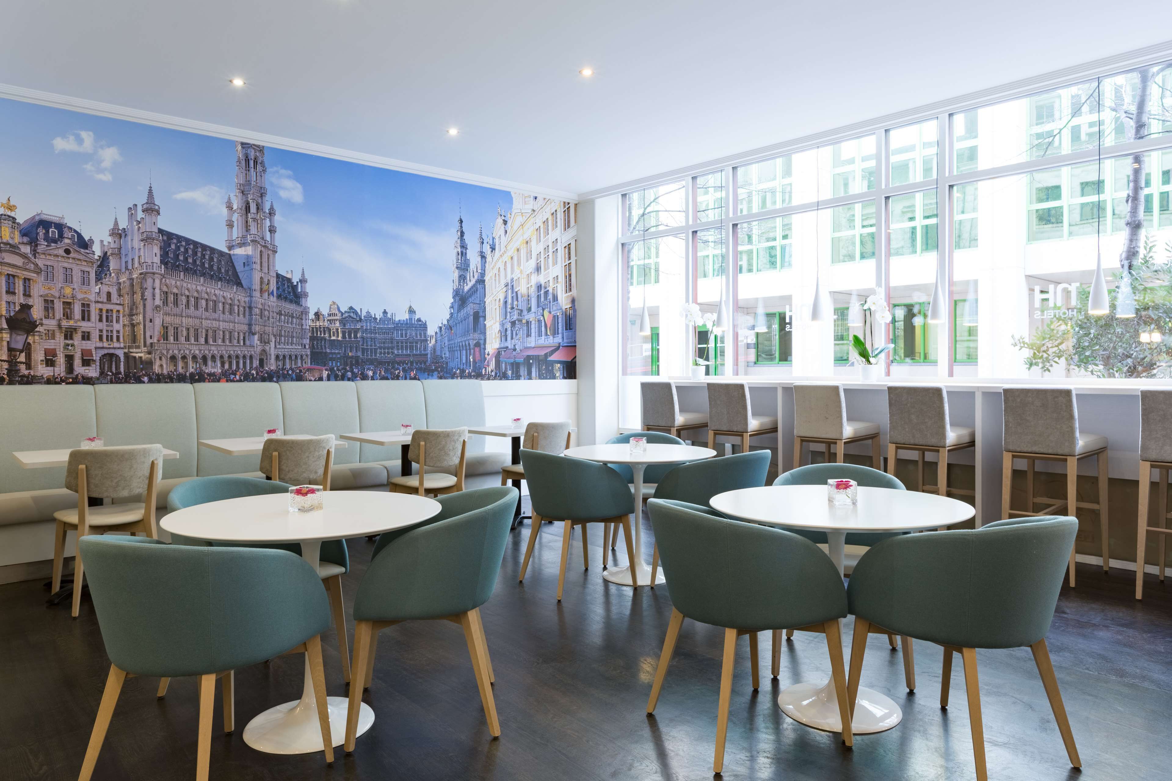 Un comedor moderno con mesas redondas y sillas color turquesa, un gran mural de un paisaje urbano y ventanas amplias que ofrecen vistas de edificios.