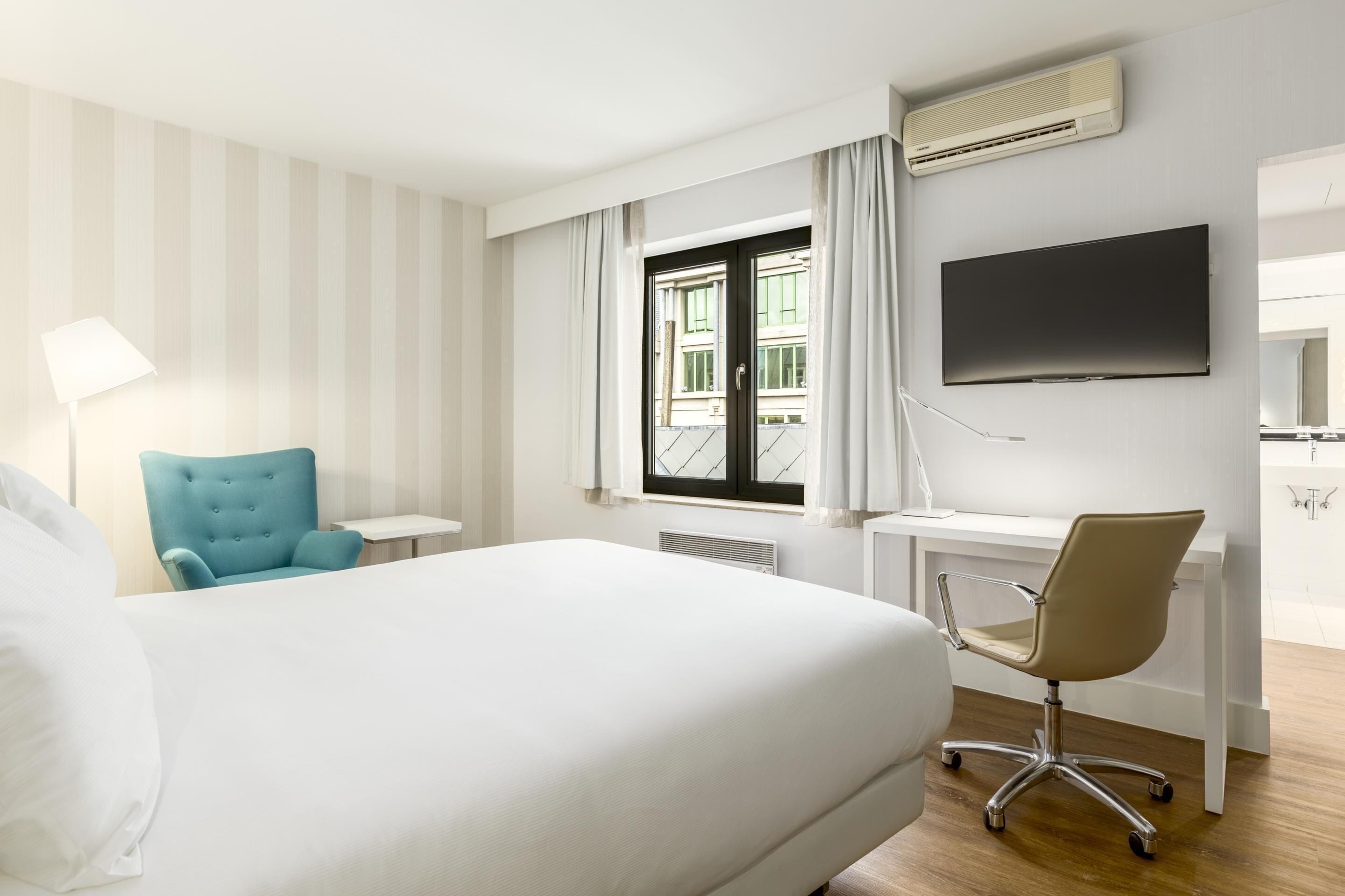 A hotel room with a bed, a desk, a chair, a TV, and a window with curtains.