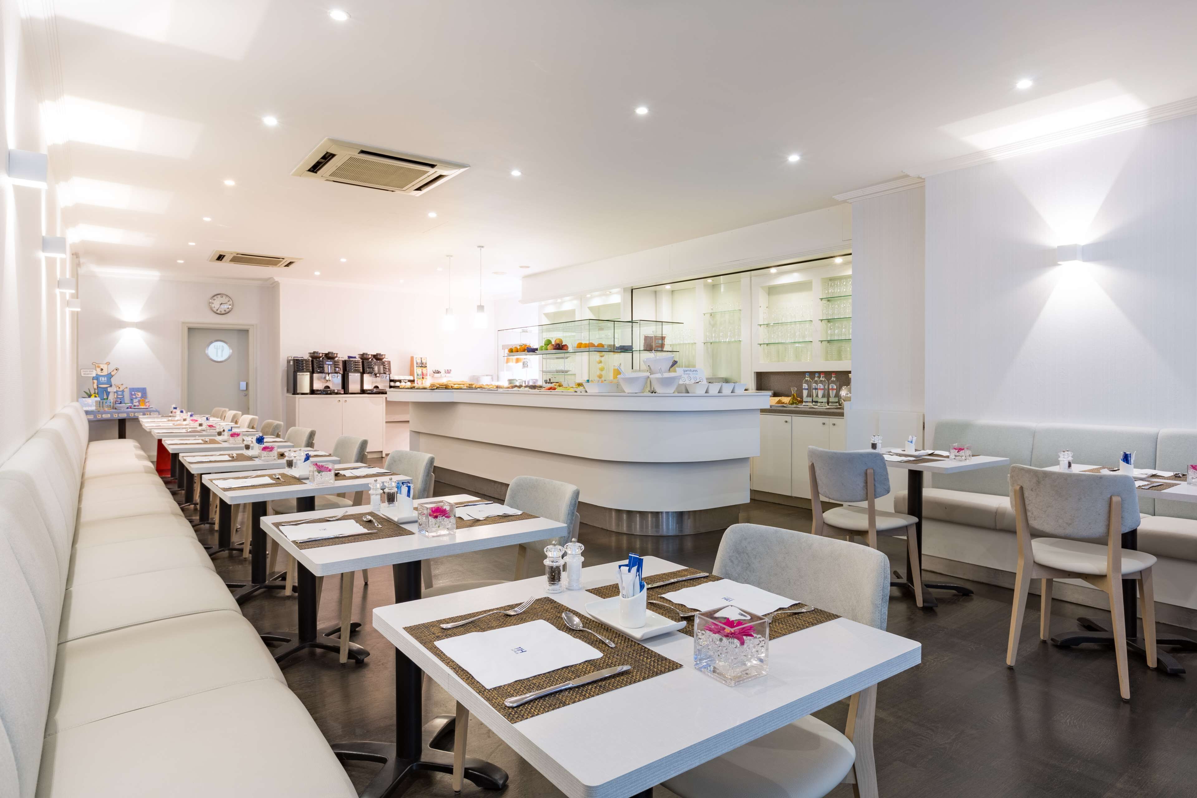 A modern restaurant with white tables, grey chairs, and a bar area.