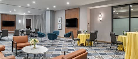 Un vestíbulo de hotel con piso de baldosas, varias zonas de estar, mesas con manteles amarillos y un televisor de pantalla plana grande montado en la pared.