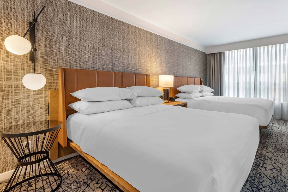 A hotel room with two beds, a small table, a wall-mounted lamp, and a large window with curtains.