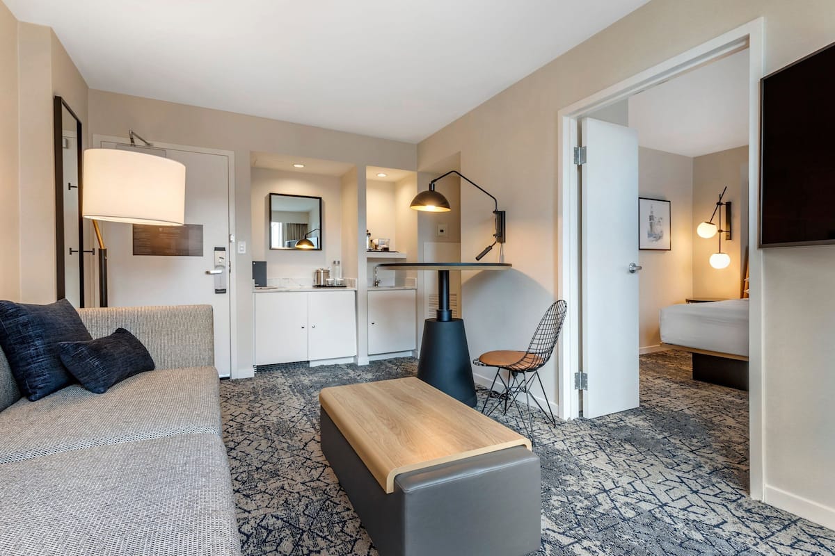 A modern hotel room with a sofa, a wooden bench, a dining area, and a separate sleeping area.