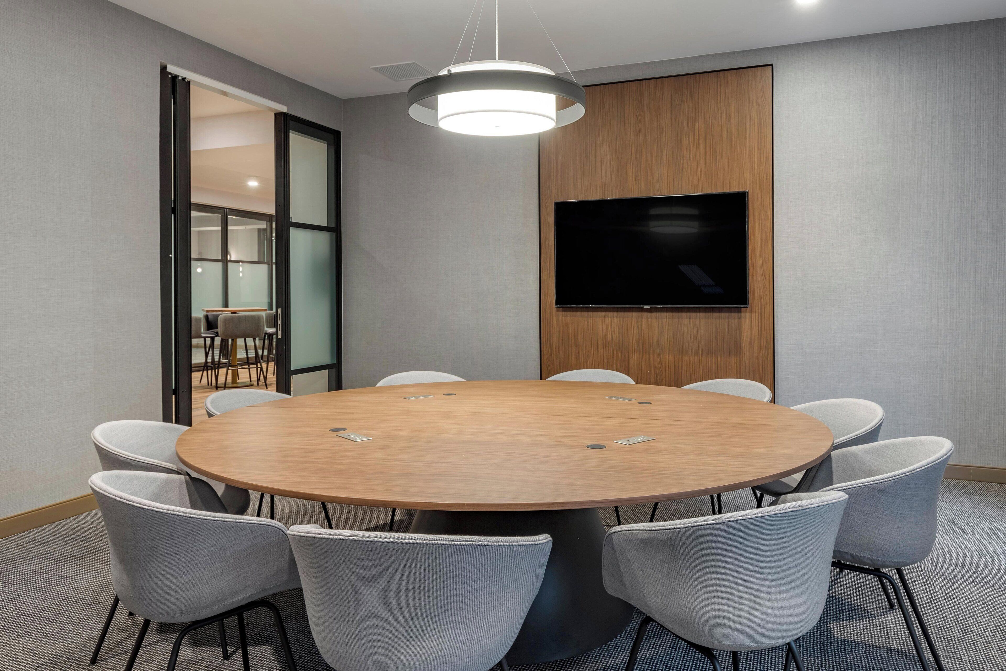 A conference room with a round table, eight chairs, a flat-screen TV, and a wooden accent wall.