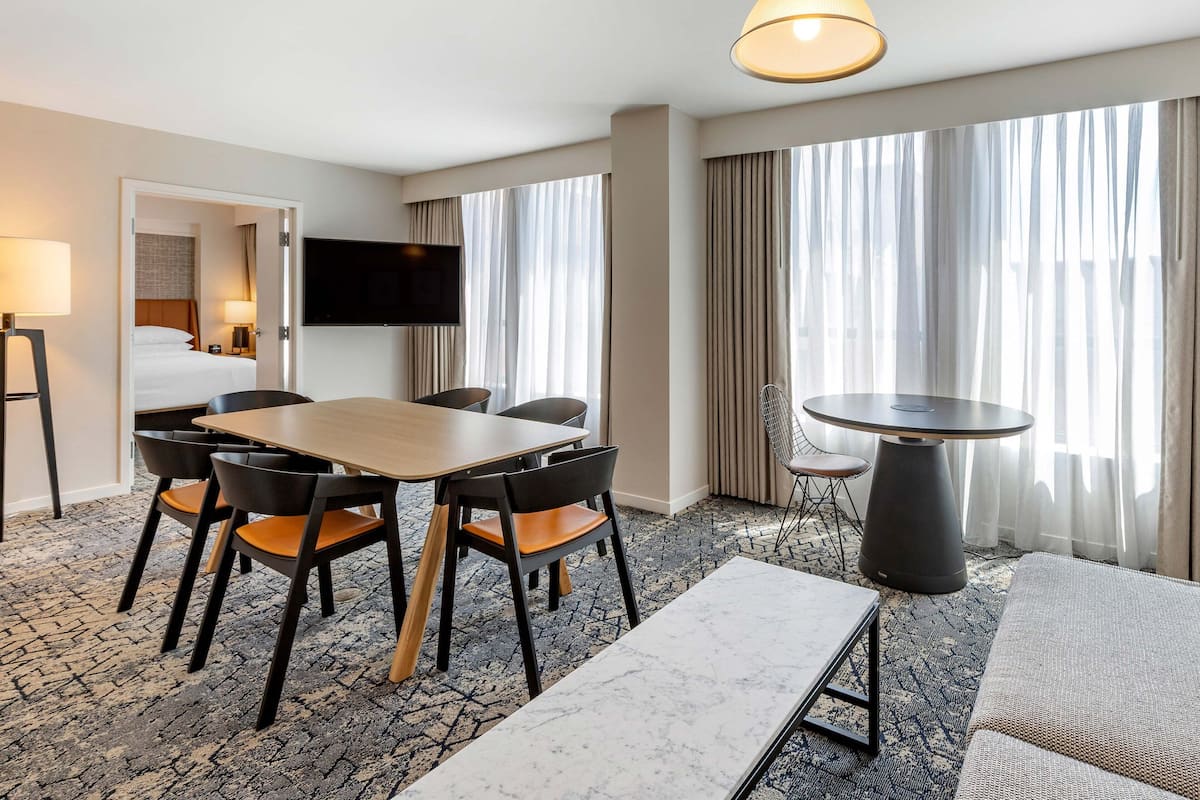 A hotel room with a dining area, a TV mounted on the wall, a large window with curtains, and a carpeted floor.