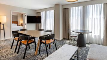 A hotel room with a dining area, a TV mounted on the wall, a large window with curtains, and a carpeted floor.