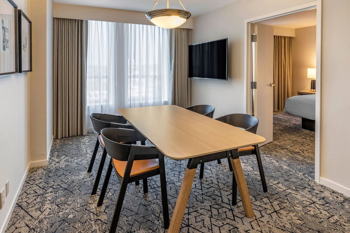 A hotel room with a large bed, a TV mounted on the wall, a dining table with chairs, and a carpeted floor.
