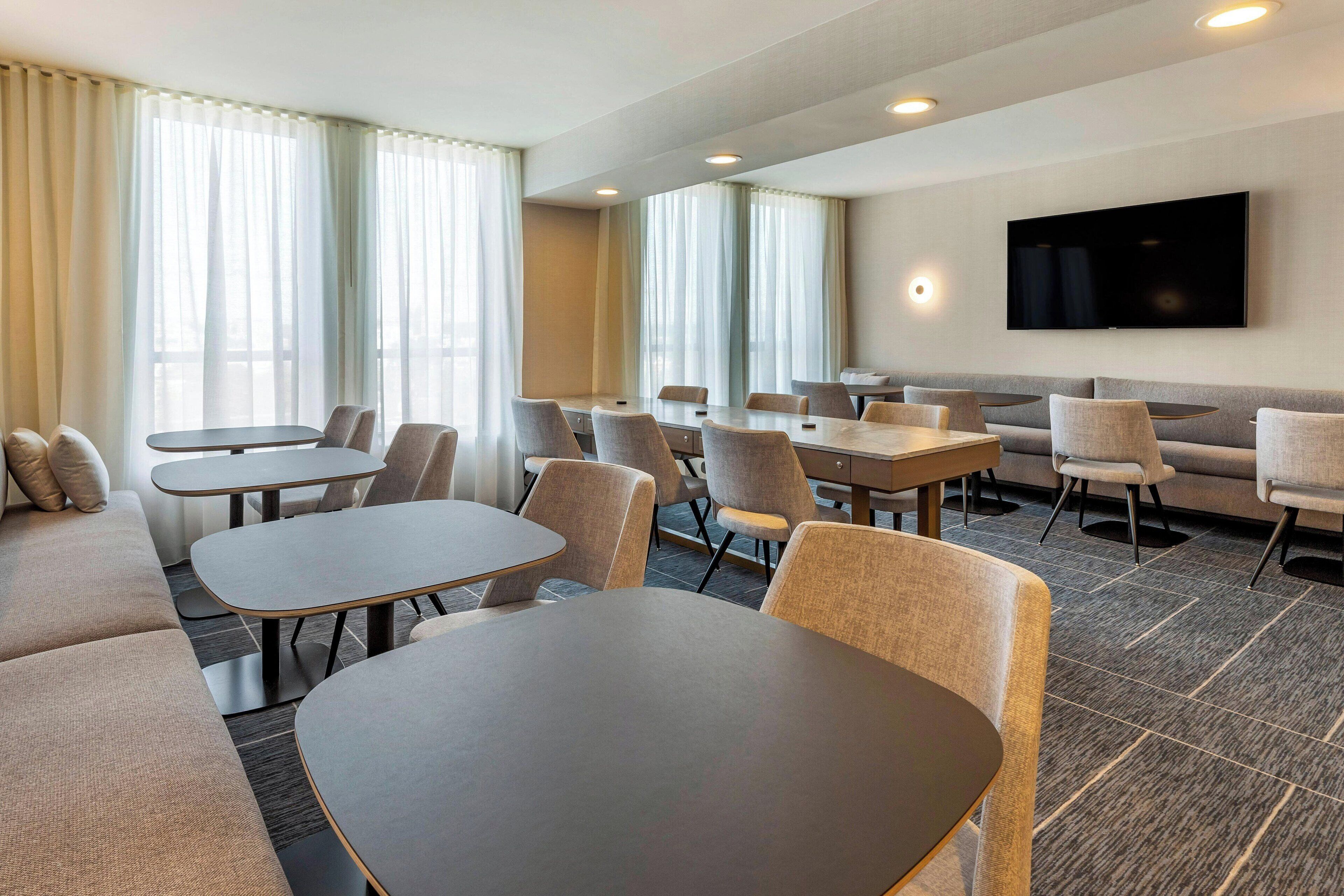 Une salle de conférence d’hôtel équipée de tables et de chaises, d’un canapé, d’un téléviseur à écran plat et de grandes fenêtres avec des rideaux.