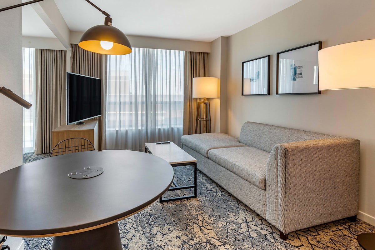A hotel room with a round table, a sofa, a TV, a lamp, and framed pictures on the wall.