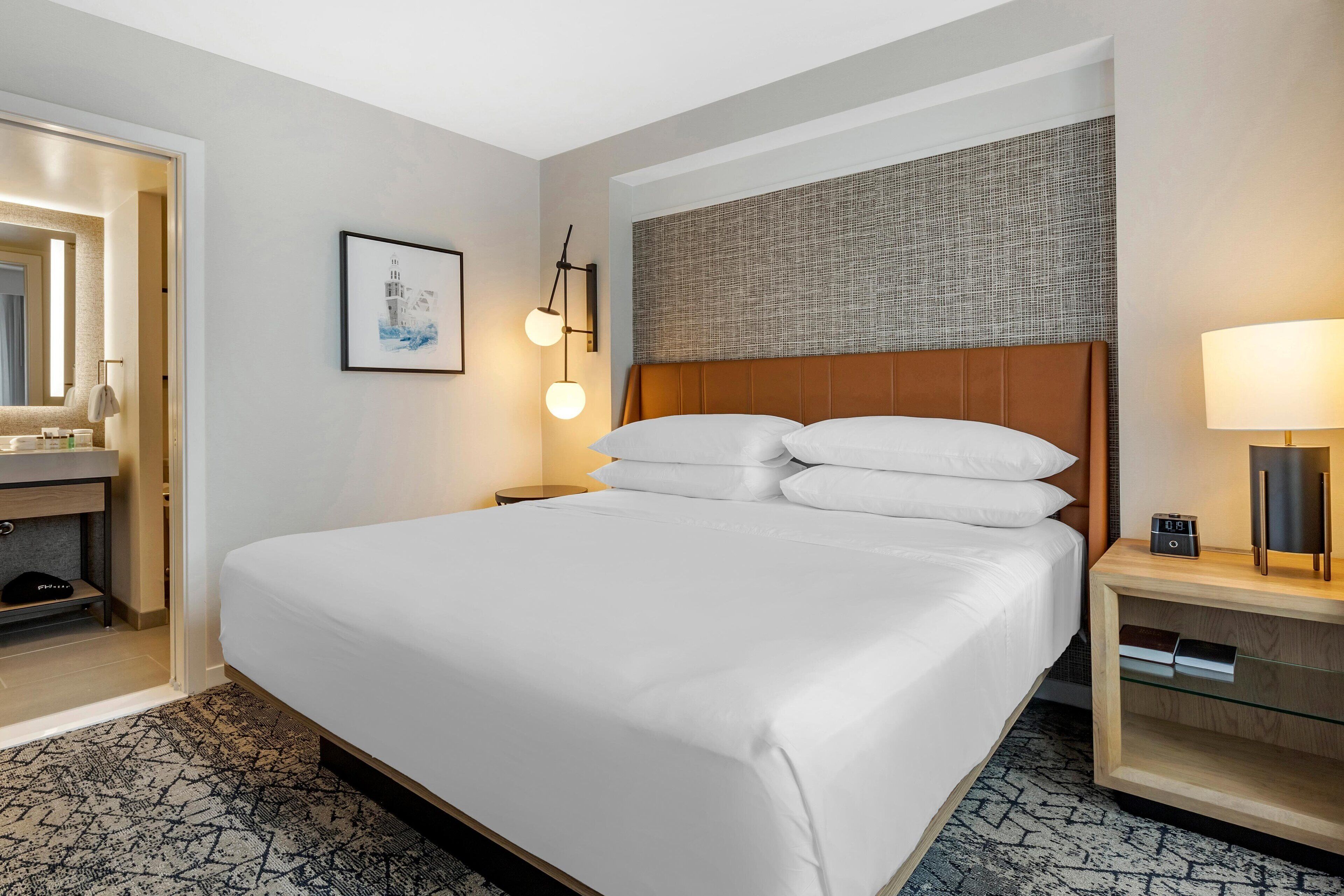 Una habitación de hotel con una cama grande, una mesita de noche con lámpara, una pequeña mesita auxiliar, un espejo y un cuadro en la pared.