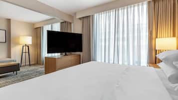 Ein Hotelzimmer mit einem großen Bett, einem Flachbildfernseher, einem hölzernen Schminktisch, einer Schreibtischlampe und einem großen Fenster mit Vorhängen.