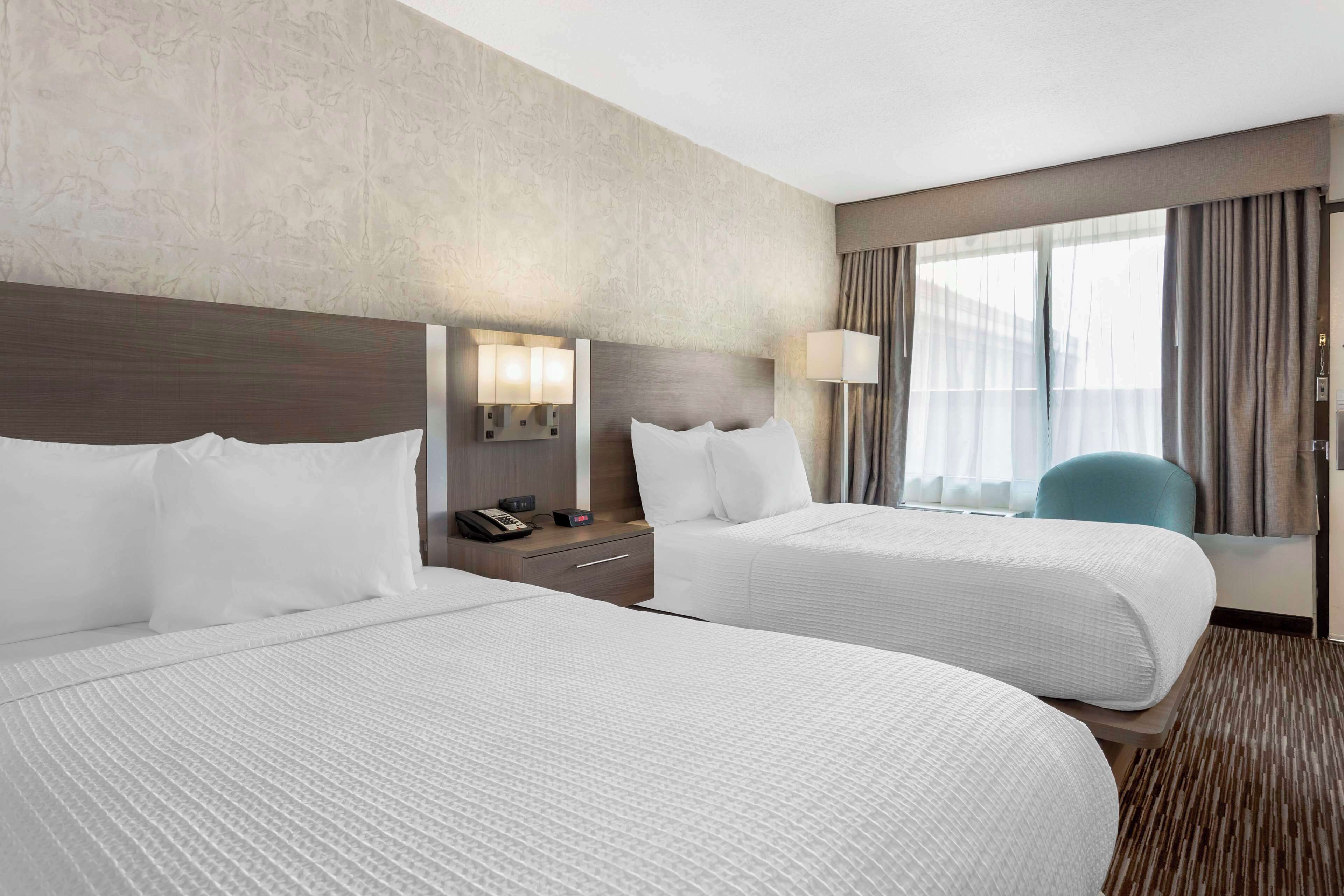 A hotel room with two beds, a nightstand with a phone and lamp, and a view of the city through large windows.