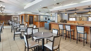 Un amplio comedor de hotel con varias mesas redondas y taburetes de bar, una zona de cocina y una pared decorativa de piedra.