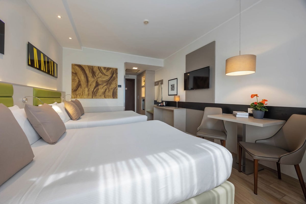 A hotel room with a large bed, a desk with a chair, a TV, and artwork on the walls.