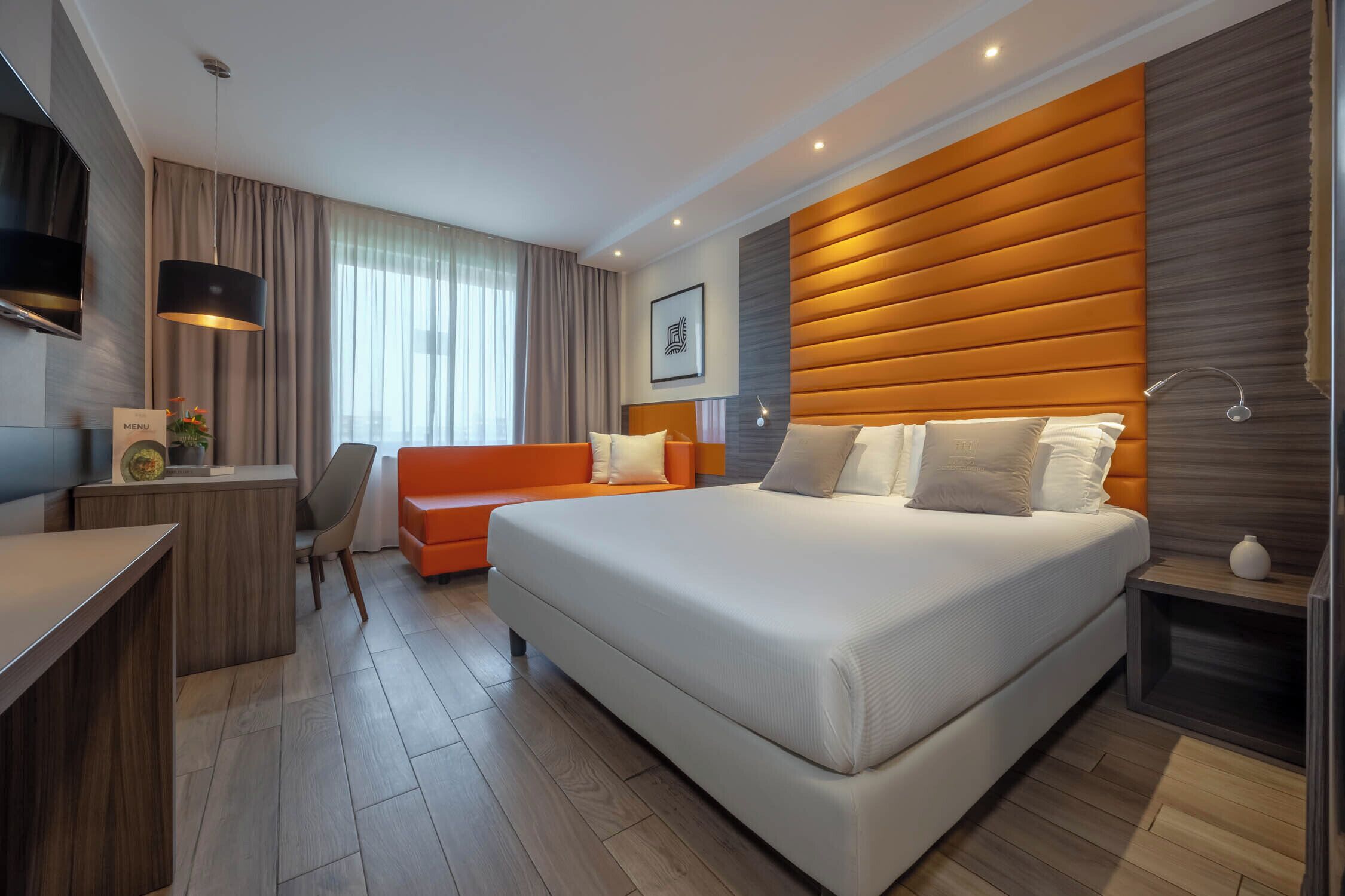A modern hotel room with a large bed, a desk, a chair, and a sofa.