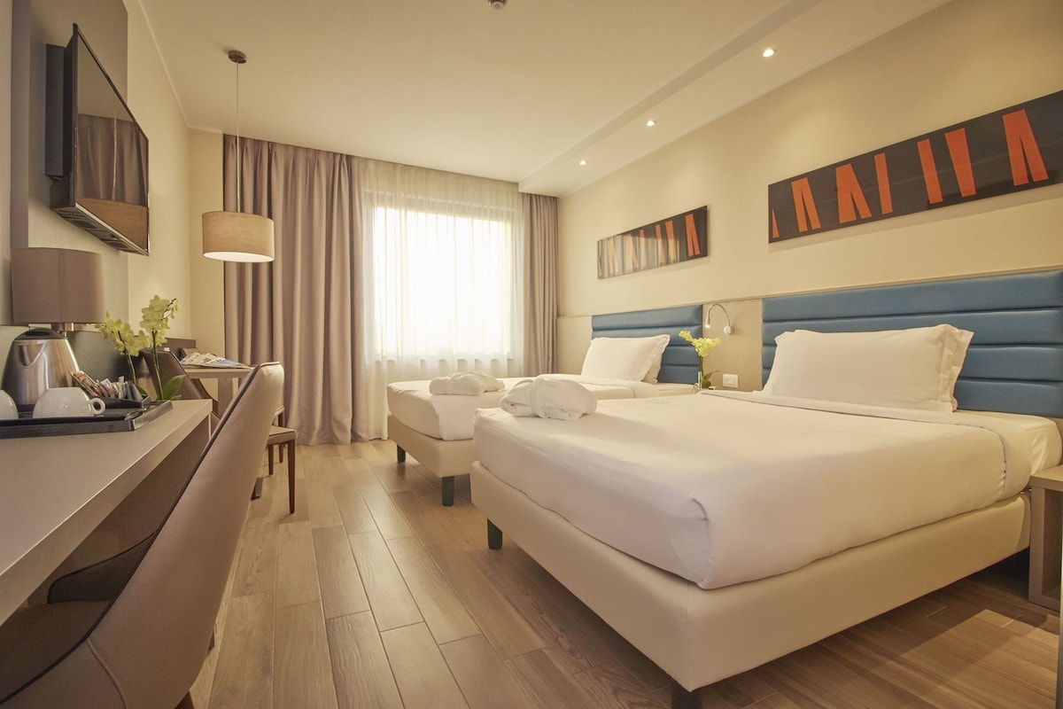 A hotel room with a large bed, a desk with a lamp, a chair, and a window with curtains.