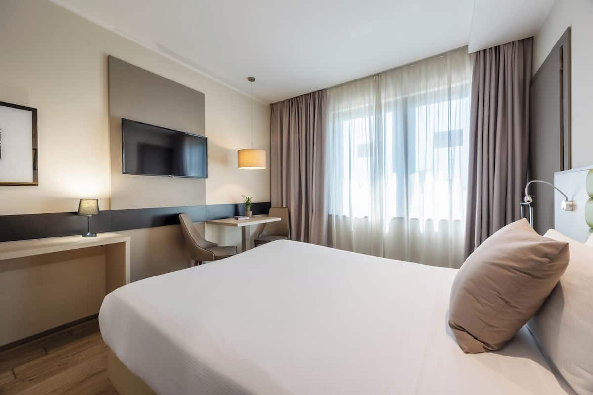 A hotel room with a bed, a desk, a chair, a TV, and a window with curtains.