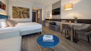 A modern hotel room with a bed, a desk, a chair, a TV, and a painting on the wall.