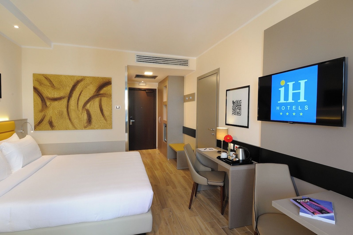 A hotel room with a bed, desk, chair, TV, and artwork.