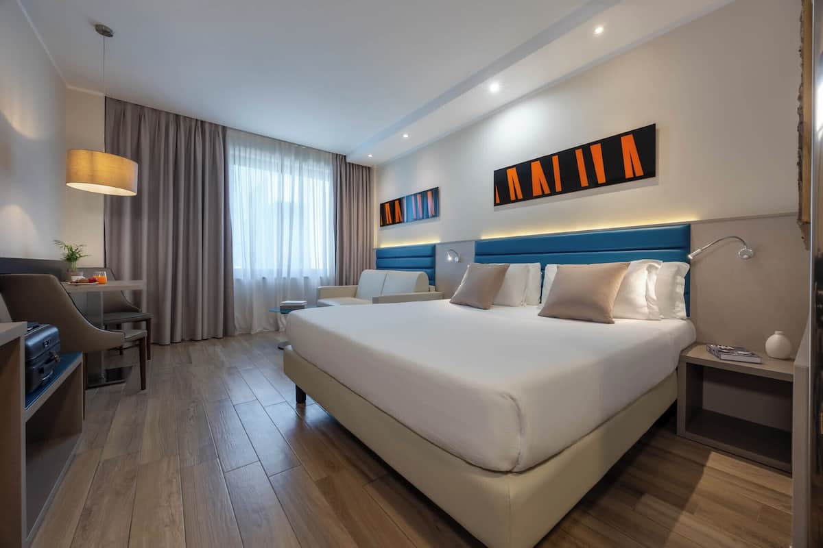 A modern hotel room with a large bed, a desk, a chair, a lamp, and a large window with curtains.